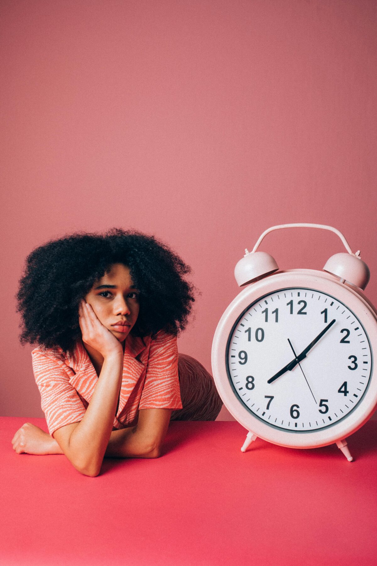A young woman lying down and propping her head up, next to a giant pink alarm clock.