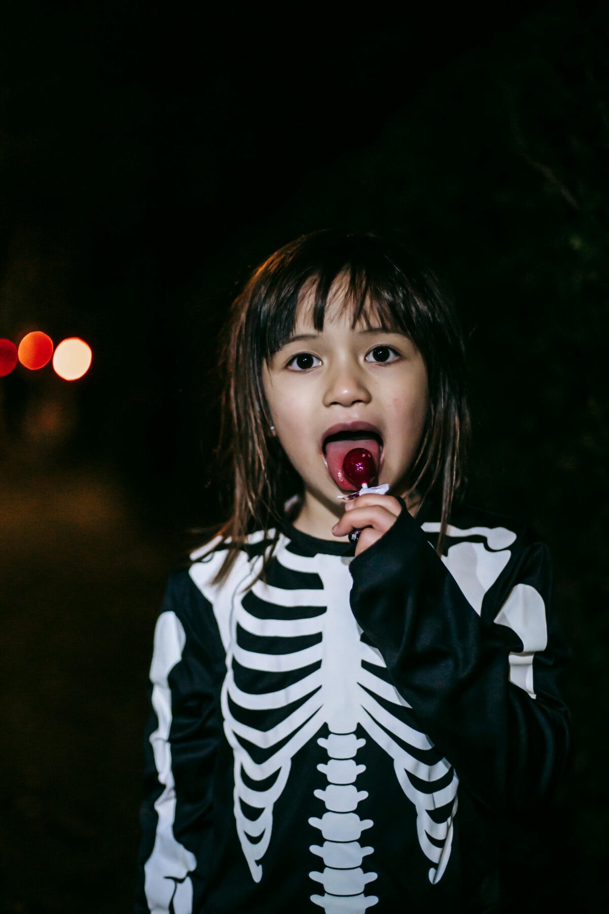 Young girl in skeleton costumer sucking on a lollypop.