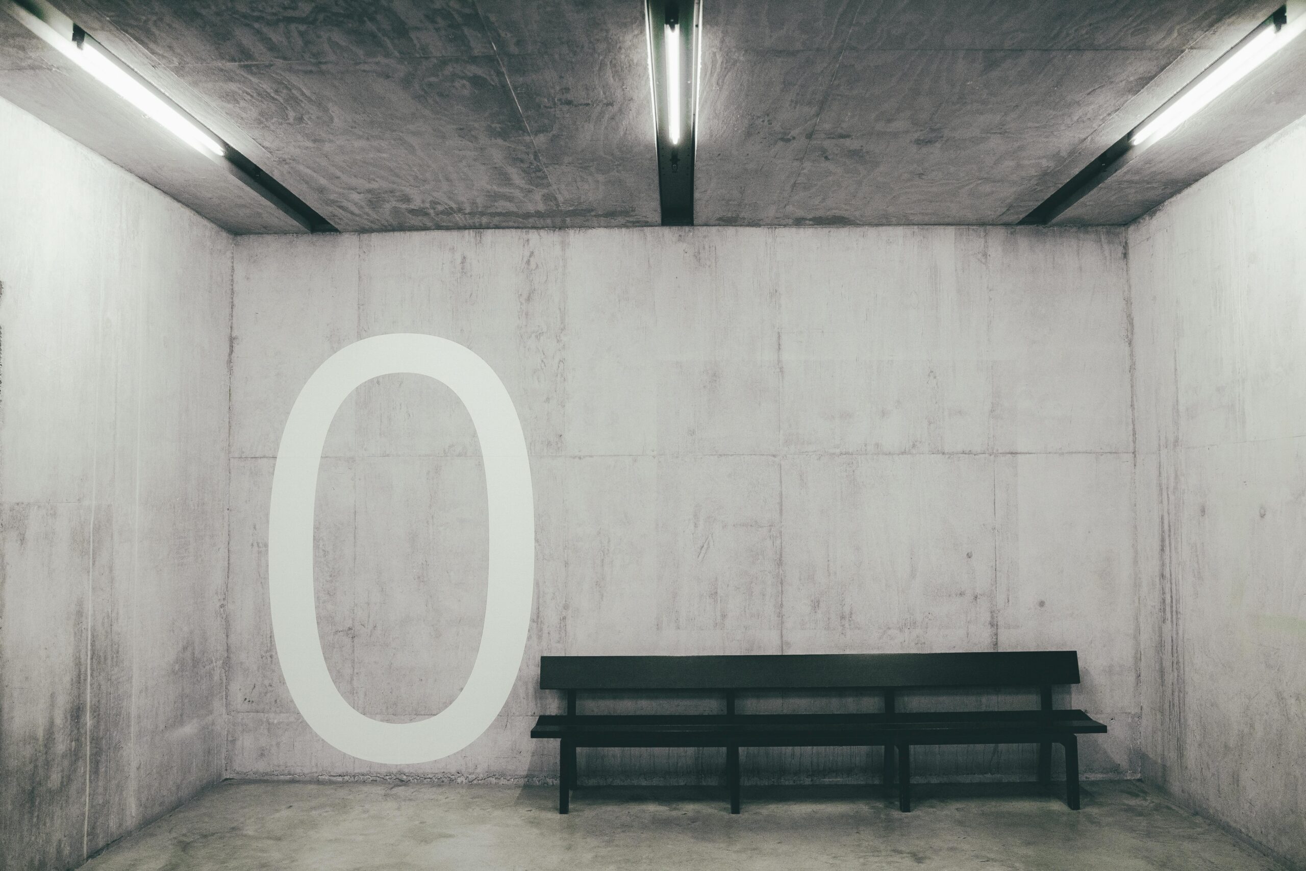 A bench in an austere room with a Zero drawn on the wall.