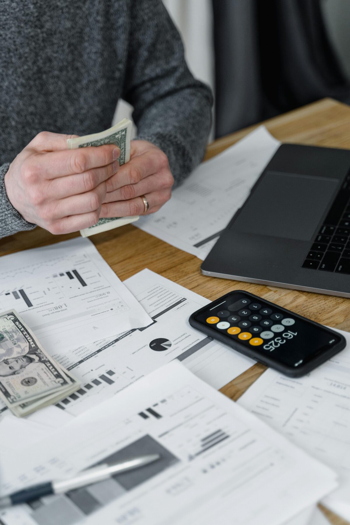 Hands of a person holding cash while sitting at a desk with a calculator, computer and papers.